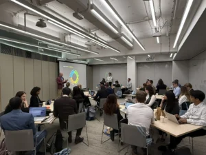 Facilitator presenting the NASA 4-D "Four Dimensions" model (mission-building, people-building, team-building, system-building) to participants seated at tables with laptops in a training room